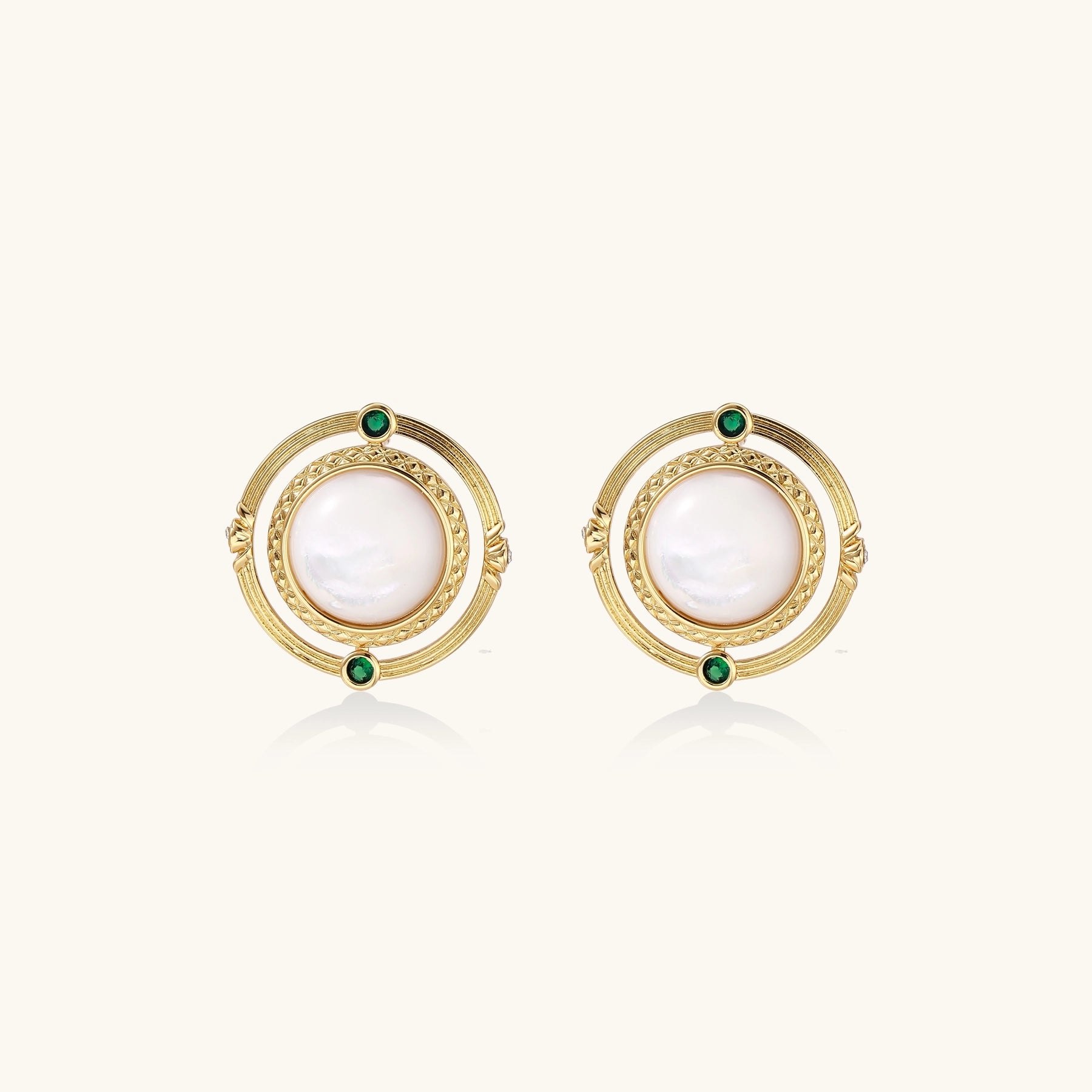 Close-up of gold-plated circular earrings with white mother-of-pearl centers and green zirconia accents on cream background.