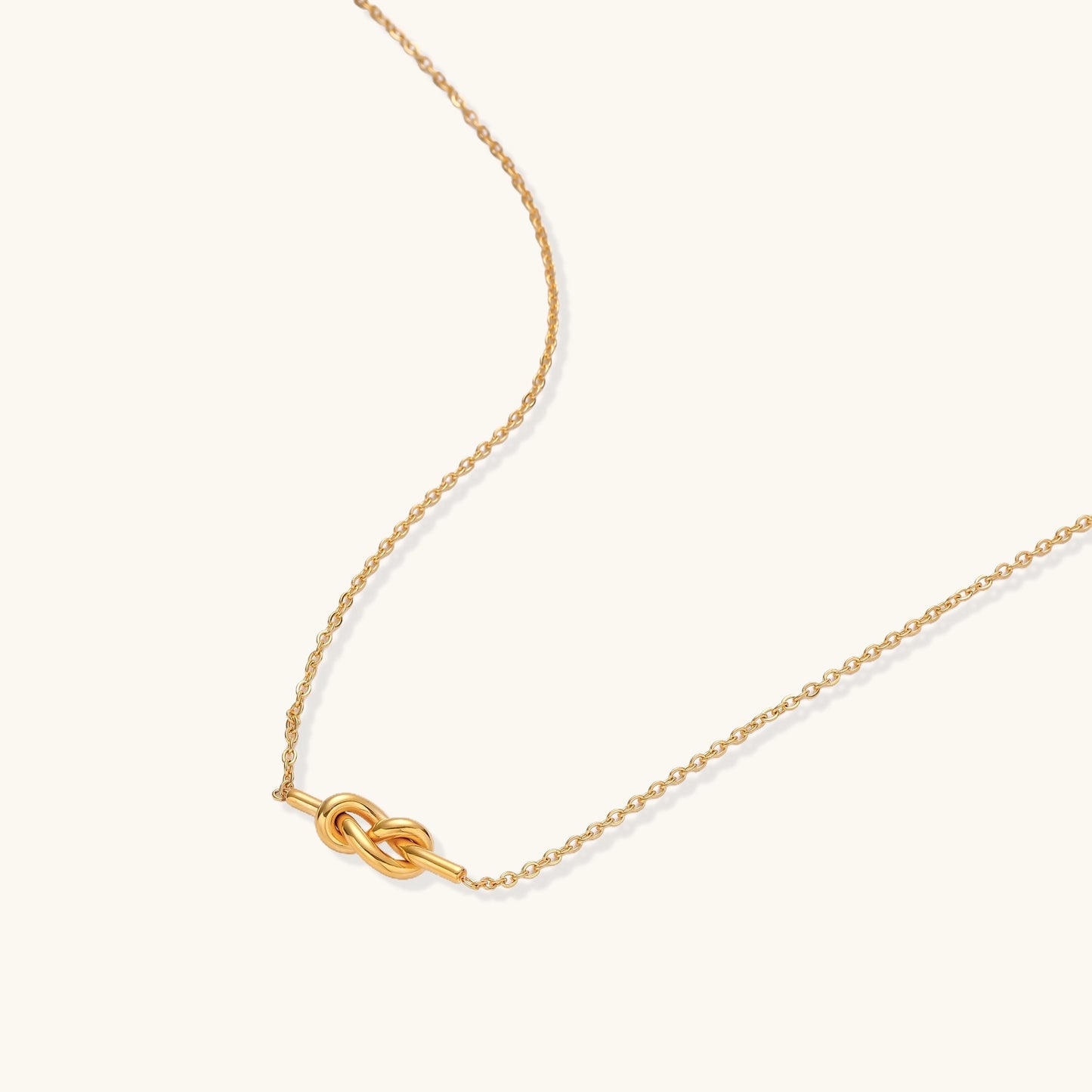 Dainty 18K gold plated stainless steel chain necklace with minimalist knot pendant on cream background.